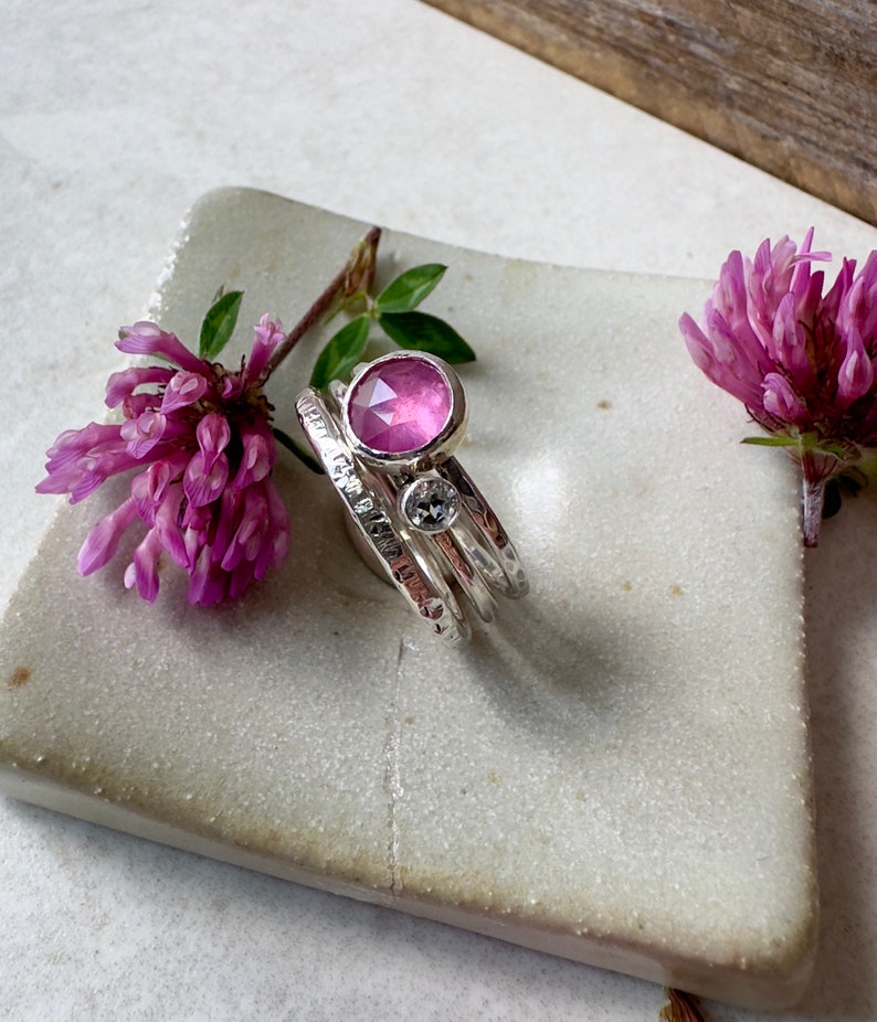 K&ouml;nnte beinhalten: Drei &uuml;bereinander gestapelte Silberringe, der oberste Ring hat einen rosafarbenen Edelstein, der mittlere Ring einen kleineren, klaren Edelstein und der unterste Ring ist ein schlichtes Band.