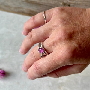 K&ouml;nnte beinhalten: Ein silberner Ring mit einem rosafarbenen Edelstein in der Mitte. Der Ring wird an einem Finger mit zwei weiteren silbernen B&auml;ndern getragen.