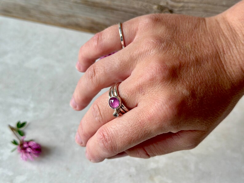 K&ouml;nnte beinhalten: Ein silberner Ring mit einem rosafarbenen Edelstein in der Mitte. Der Ring wird an einem Finger mit zwei weiteren silbernen B&auml;ndern getragen.