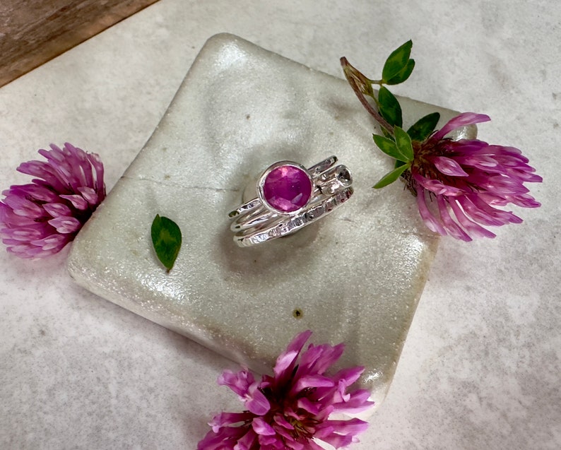 K&ouml;nnte beinhalten: Ein silberner Ring mit einem rosafarbenen Edelstein, der in einem strukturierten Band gefasst ist. Der Ring ist von rosa Kleebl&uuml;ten umgeben.