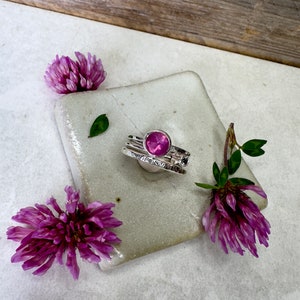 May include: A set of three silver rings with a pink gemstone in the centre ring. The rings are on a white ceramic tile with pink flowers on either side.