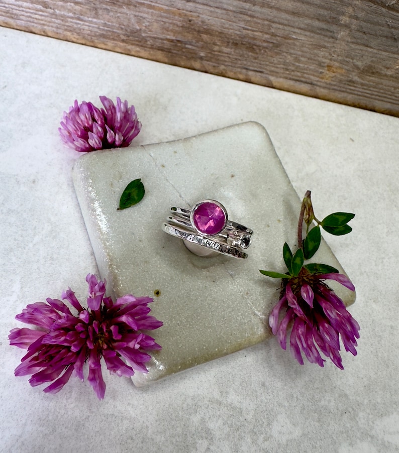 K&ouml;nnte beinhalten: Ein Satz aus drei Silberringen mit einem rosafarbenen Edelstein im mittleren Ring. Die Ringe liegen auf einer wei&szlig;en Keramikfliese mit rosa Blumen auf beiden Seiten.