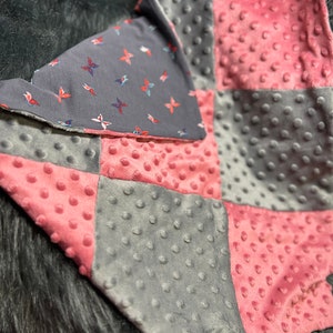 May include: A patchwork baby blanket with a gray and pink color scheme. The blanket is made of soft, textured fabric and features a variety of patterns, including polka dots and butterflies.