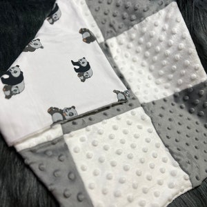 May include: A white and grey baby blanket with a panda bear print. The blanket is made of soft, textured fabric.