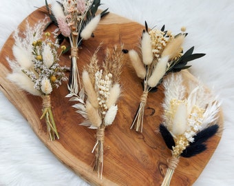 May include: Five small bouquets of dried flowers arranged on a wooden surface. The bouquets feature a variety of dried grasses, pampas grass, and white and cream colored flowers.
