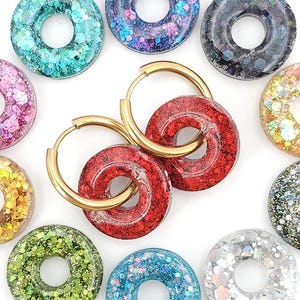 Può includere: Orecchini a cerchio dorati con un ciondolo a forma di ciambella rossa glitterata. Intorno agli orecchini ci sono altri ciondoli a forma di ciambella in vari colori, tra cui turchese, viola, verde e argento, tutti con dettagli glitterati. Sfondo bianco.