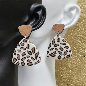 May include: A pair of earrings featuring a wooden top and a triangular pendant with a white base and brown animal print design. The earrings are hanging from a black and white ear-shaped display.