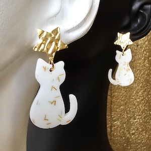 Pode incluir: Um par de brincos em forma de gato. Os brincos apresentam uma silhueta de gato branco com manchas douradas, suspensa de um pino de estrela dourada texturizada. Os brincos são projetados para serem usados na orelha.