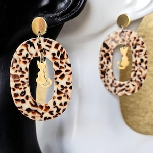 Puede incluir: Pendientes ovalados con estampado carey y silueta de gato dorada. Los pendientes tienen un disco dorado en la parte superior y un dije de gato dorado colgando en el centro. Fondo blanco y negro.