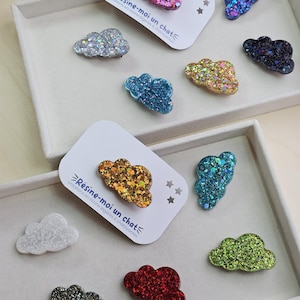 Op de afbeelding: Een verzameling glitterende, wolk-vormige harsaccessoires in verschillende kleuren, waaronder paars, zilver, blauw, goud en groen. Sommige worden gepresenteerd op witte kaarten met tekst. De accessoires zijn gerangschikt in een bakje.