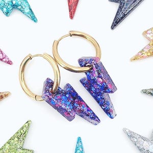 Könnte beinhalten: Goldene Creolen-Ohrringe mit lila und blauen Glitzer-Blitz-Anhängern. Zusätzliche farbenfrohe Glitzer-Blitz-Anhänger in verschiedenen Farben sind um die Ohrringe herum verstreut.