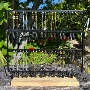 May include: A display of various ear cuff earrings with dangling chains. The earrings feature different charms, including moons, swords, stars, and spikes. The earrings are displayed on a black stand with a wooden base.