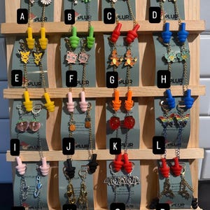 May include: A display of earplugs with decorative charms. Styles include cartoon characters, hearts, and flowers. The earplugs are in various colors, including yellow, green, red, and blue, and are displayed on a wooden rack.
