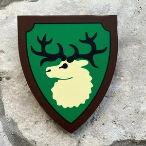 May include: A wooden shield-shaped emblem with a green background featuring a cream-colored deer head and black antlers. The shield is framed by a brown border and is set against a stone surface.