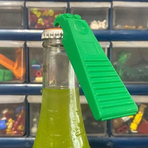 May include: A green plastic bottle opener with a textured surface and a small, round button on the top. The opener is being used to open a green glass bottle.
