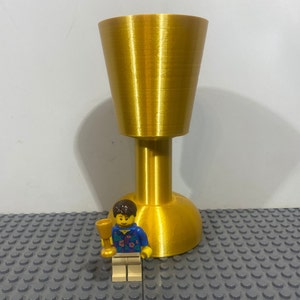 Pode incluir: Um troféu dourado, impresso em 3D, com uma base larga e acampanada e um caule cilíndrico alto. Uma pequena figura de Lego amarela está em frente ao troféu.