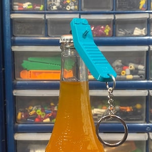 May include: A blue bottle opener keychain attached to a glass bottle of orange juice. The bottle opener has a textured surface and a silver key ring.