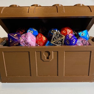May include: A brown plastic treasure chest with a hinged lid is open and filled with a variety of colorful dice. The dice are made of different materials, including plastic, metal, and glass. The dice are in various colors, including red, blue, green, and purple.