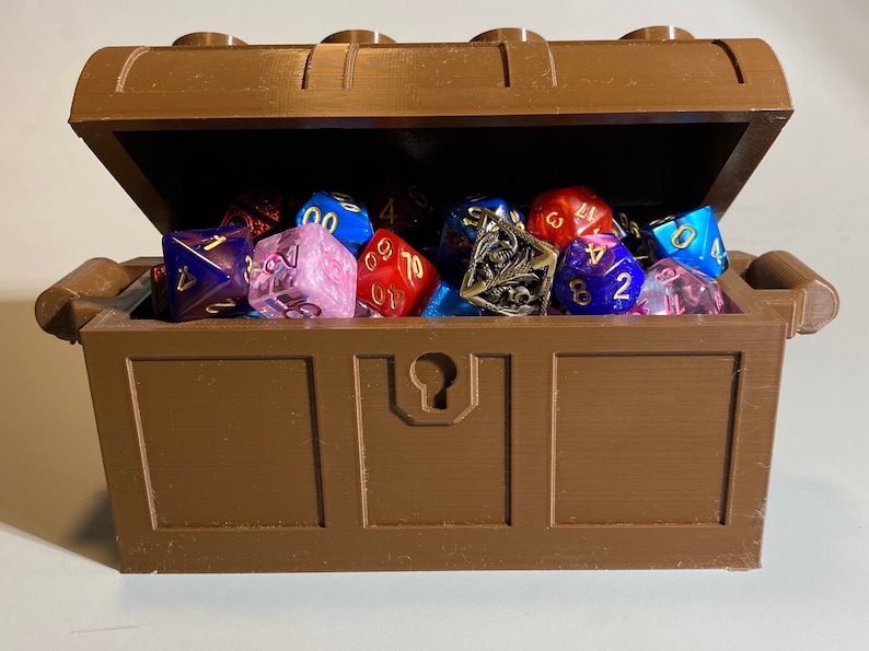 May include: A brown plastic treasure chest with a hinged lid is open and filled with a variety of colorful dice. The dice are made of different materials, including plastic, metal, and glass. The dice are in various colors, including red, blue, green, and purple.