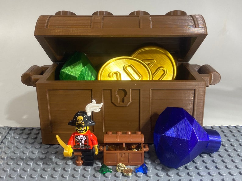 May include: A brown wooden treasure chest with a hinged lid is open, revealing a green and blue gem, gold coins, and a smaller brown wooden chest. A pirate Lego minifigure stands in front of the chest, holding a sword.