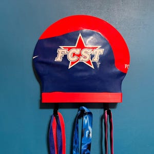 May include: A wall-mounted swim cap holder in red and navy blue, featuring the text "FCST" inside a white star. The holder has three hooks with colorful ribbons hanging from them. The background is a solid blue wall.