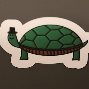 May include: A green turtle wearing a black top hat with a white stripe. The turtle has a brown shell with a brown stripe around the edge.