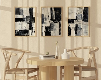 May include: Three abstract black and white paintings in wooden frames hanging on a beige wall above a wooden table with two chairs.