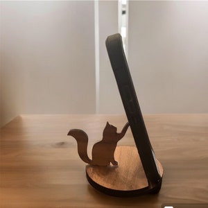 May include: A wooden phone stand featuring a squirrel silhouette. The stand is made of light and dark brown wood, with the phone resting against a dark case. The squirrel appears to be holding the phone in place. The stand is approximately 4 inches long.
