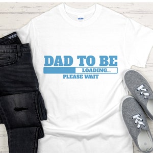May include: White t-shirt with blue text that reads "Dad to be... Loading... Please wait". The text is displayed above a blue progress bar.