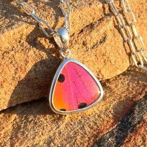 Könnte beinhalten: Eine silberne Halskette mit einem dreieckigen Anhänger. Der Anhänger hat ein leuchtend rosa-oranges Design mit schwarzen Punkten, eingefasst in einen silbernen Rahmen. Die Halskette wird auf einer strukturierten, braunen Oberfläche präsentiert.