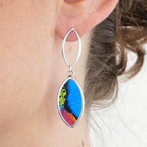 Puede incluir: Pendientes colgantes plateados con un colgante ovalado. El colgante presenta un diseño vibrante en azul, verde y rojo, posiblemente un ala de mariposa. Los pendientes cuelgan de un óvalo más pequeño y alargado. El diseño es llamativo y colorido.