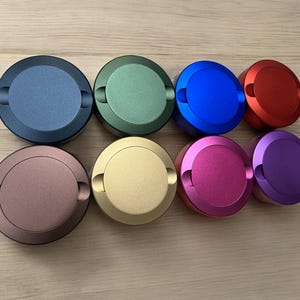 Op de afbeelding: Acht ronde metalen containers met deksels in verschillende kleuren: blauw, groen, rood, paars, goud, bruin, roze en blauw. De containers zijn in een halve cirkel op een houten oppervlak gerangschikt.