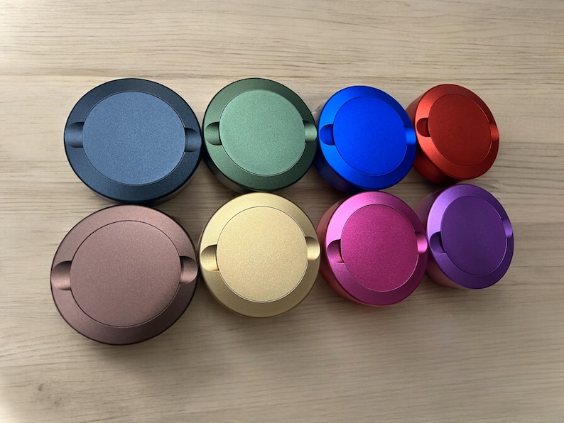 Op de afbeelding: Acht ronde metalen containers met deksels in verschillende kleuren: blauw, groen, rood, paars, goud, bruin, roze en blauw. De containers zijn in een halve cirkel op een houten oppervlak gerangschikt.