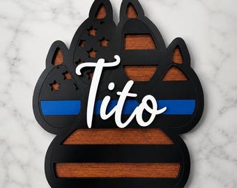 Può includere: Una decorazione da parete in legno a forma di zampa con cornice nera, una sottile linea blu e intagli a forma di stella. Le venature del legno naturale sono visibili attraverso gli intagli e la scritta Tito è applicata in bianco al centro.