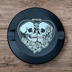 Op de afbeelding: Rond zwart auto-embleem met een wit ontwerp van twee skeletten in een hartvorm met rozen. De datum 10/31/22 is boven de skeletten gedrukt.