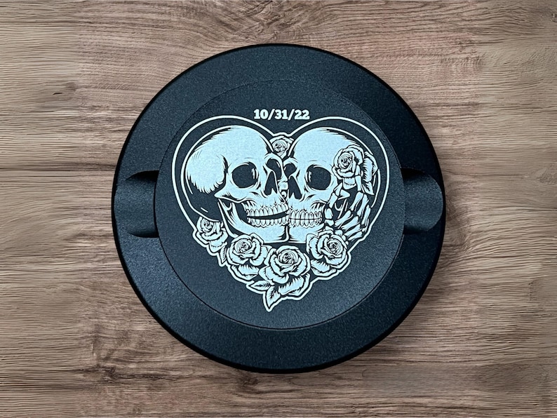 Op de afbeelding: Rond zwart auto-embleem met een wit ontwerp van twee skeletten in een hartvorm met rozen. De datum 10/31/22 is boven de skeletten gedrukt.