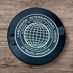 Op de afbeelding: Een zwart, rond embleem met witte tekst en een globe-ontwerp. De tekst luidt "Laborers' International Union of North America" met de woorden "Rechtvaardigheid, Eer, Kracht" rond de globe. De tekst bevat ook "Georganiseerd op 13 april 1903".