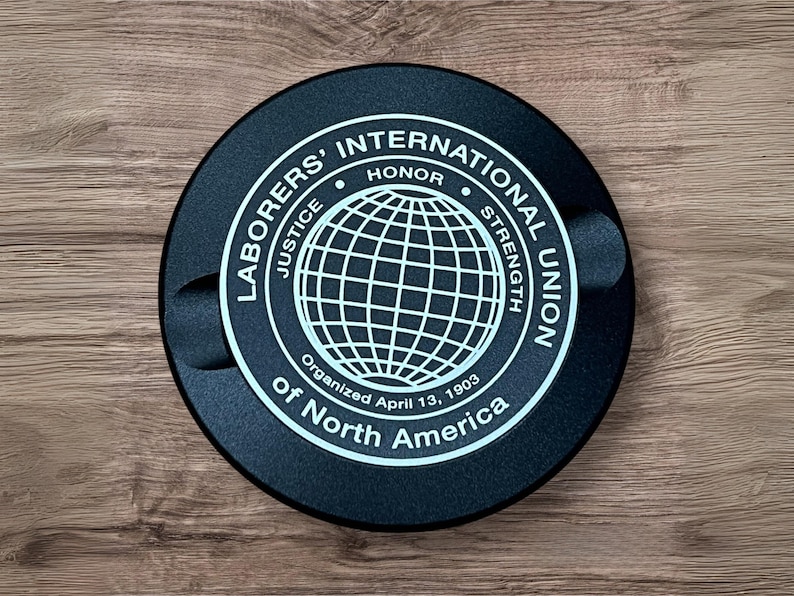 Op de afbeelding: Een zwart, rond embleem met witte tekst en een globe-ontwerp. De tekst luidt "Laborers' International Union of North America" met de woorden "Rechtvaardigheid, Eer, Kracht" rond de globe. De tekst bevat ook "Georganiseerd op 13 april 1903".