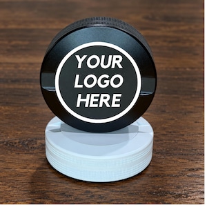 May include: A circular black grinder with a textured edge sits atop a white circular base. The center of the black grinder features a white circular border enclosing the white text "YOUR LOGO HERE" in a bold, sans-serif font, centered on the surface.