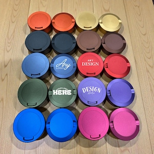 May include: An overhead shot of sixteen round, metallic containers in various colors, including orange, gold, blue, green, and purple. Some lids have text that reads "Any Logo", "Any Design", and "Your Logo Here".