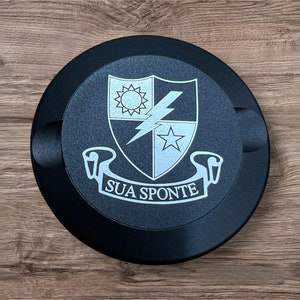 Op de afbeelding: Een zwarte ronde schijf met een wit embleem met een schild met een bliksem, een ster en een zon. Het schild wordt omringd door een wit lint met de tekst "SUA SPONTE".