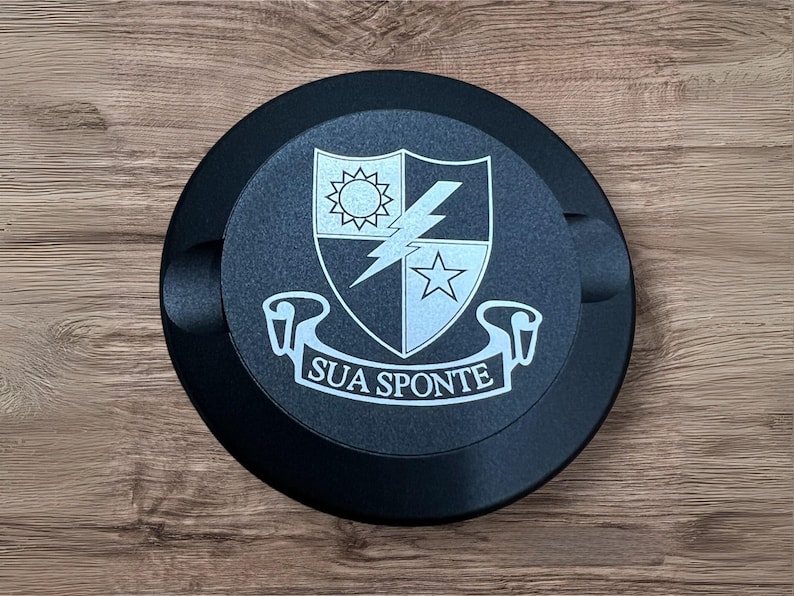 Op de afbeelding: Een zwarte ronde schijf met een wit embleem met een schild met een bliksem, een ster en een zon. Het schild wordt omringd door een wit lint met de tekst "SUA SPONTE".