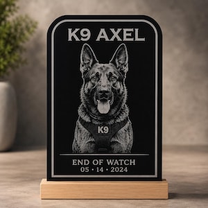 May include: A black memorial plaque with a portrait of a German Shepherd dog, the text "K9 AXEL" at the top, and "END OF WATCH 05.14.2024" at the bottom. The plaque is mounted on a wooden base.