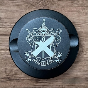 Op de afbeelding: Een rond zwart deksel met een zilveren gegraveerd wapenschild met een kruis, een herdersstaf en de tekst "AN APIZES OE".