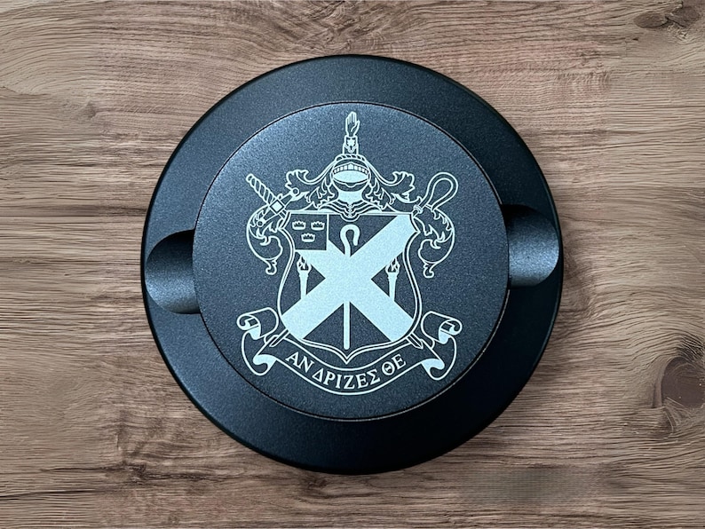 Op de afbeelding: Een rond zwart deksel met een zilveren gegraveerd wapenschild met een kruis, een herdersstaf en de tekst "AN APIZES OE".