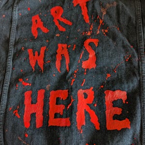 Può includere: Una giacca di jeans nera con la scritta ART WAS HERE dipinta in rosso sul retro. Le lettere presentano un effetto di vernice colante e usurata, con schizzi di vernice rossa sparsi sul tessuto scuro del capo di abbigliamento.