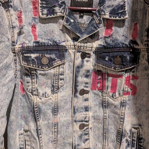 Puede incluir: Chaqueta vaquera azul claro con efecto lavado ácido desgastado. La chaqueta presenta salpicaduras de pintura rosa y la palabra "BTS" en rosa en la parte delantera. Tiene dos bolsillos en el pecho y cierre de botones.