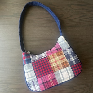 May include: A shoulder bag featuring a patchwork design composed of various plaid and checkered fabric squares in shades of red, blue, white, brown, and pink. The bag has a single dark blue strap and a zipper closure along the top edge.