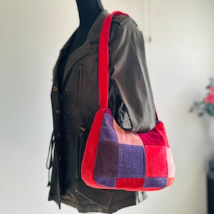 May include: A patchwork shoulder bag with a red strap. The bag features a rectangular shape with a variety of red, pink, and purple squares. The bag is displayed on a mannequin wearing a dark green jacket.