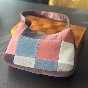 May include: A patchwork shoulder bag in shades of pink, blue, gray, and white. The bag has a curved shape with a matching strap and a dark brown base. The bag is made of a soft, textured fabric.